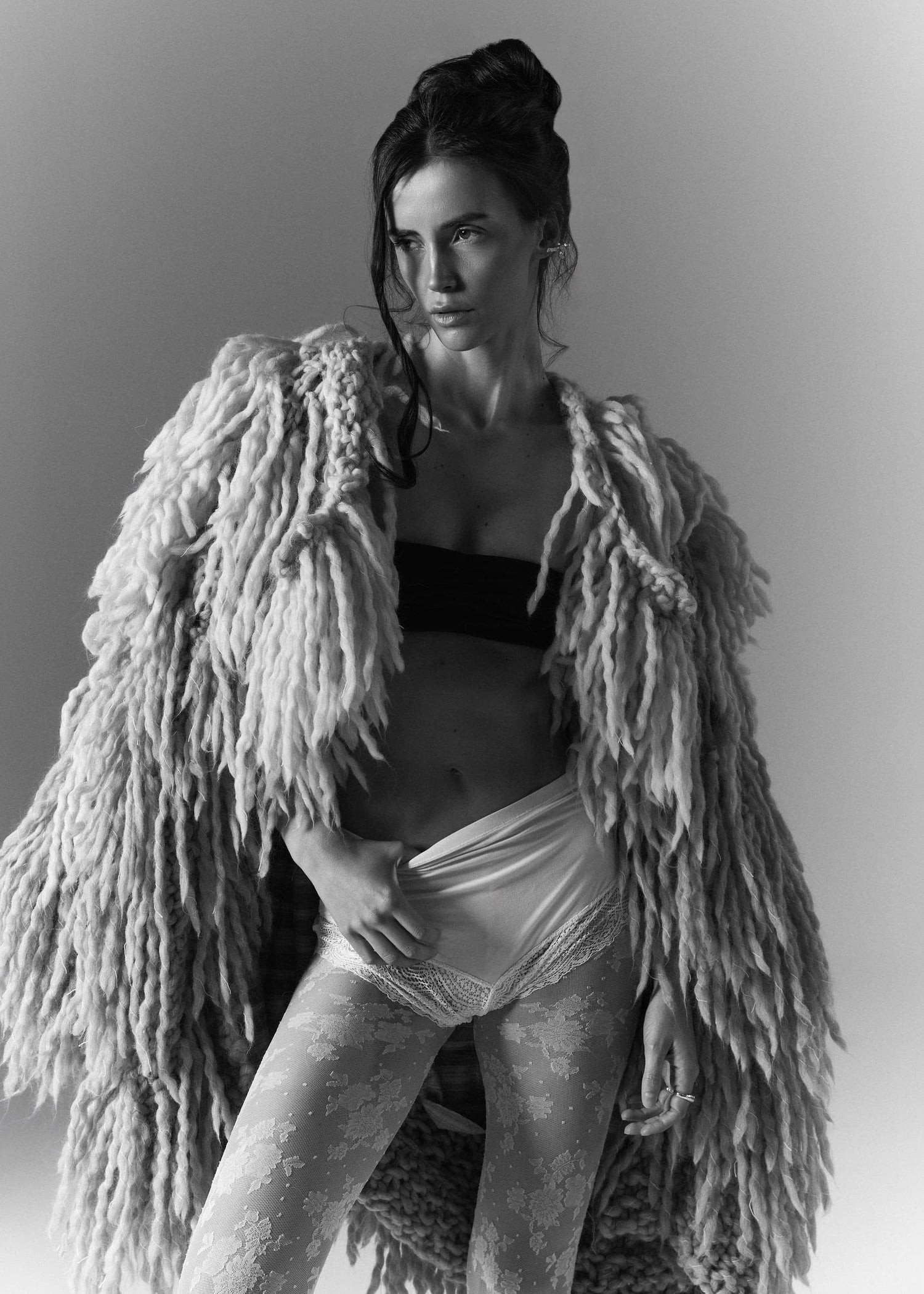 A black and white fashion portrait of a woman wearing a voluminous shaggy faux-fur jacket over a black bandeau top and white underwear, posed against a plain studio background.